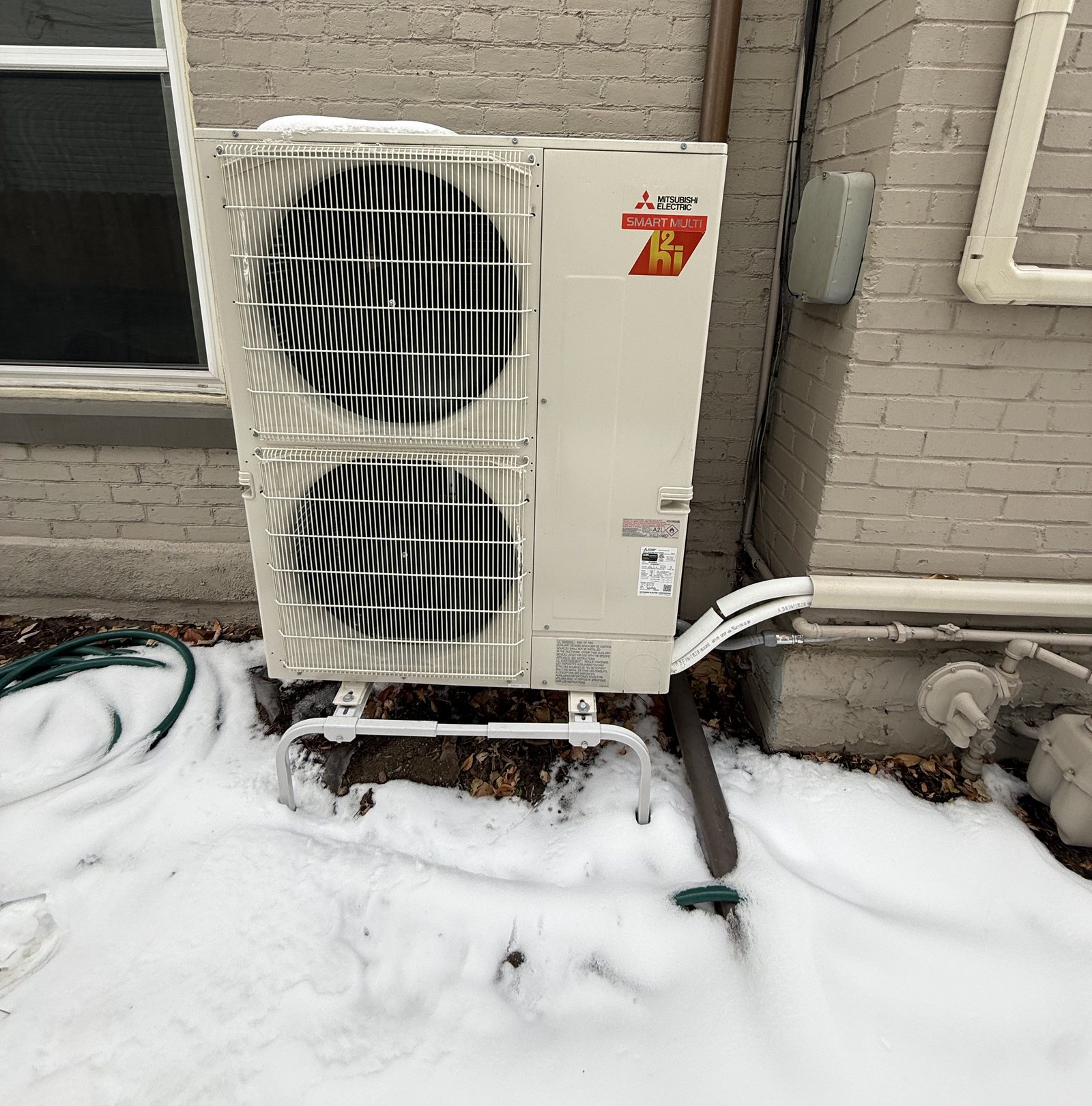 Mitsubishi Electric SMART MULTI H2i heat pump outdoor unit on a stand in fresh snow beside a Denver brick home