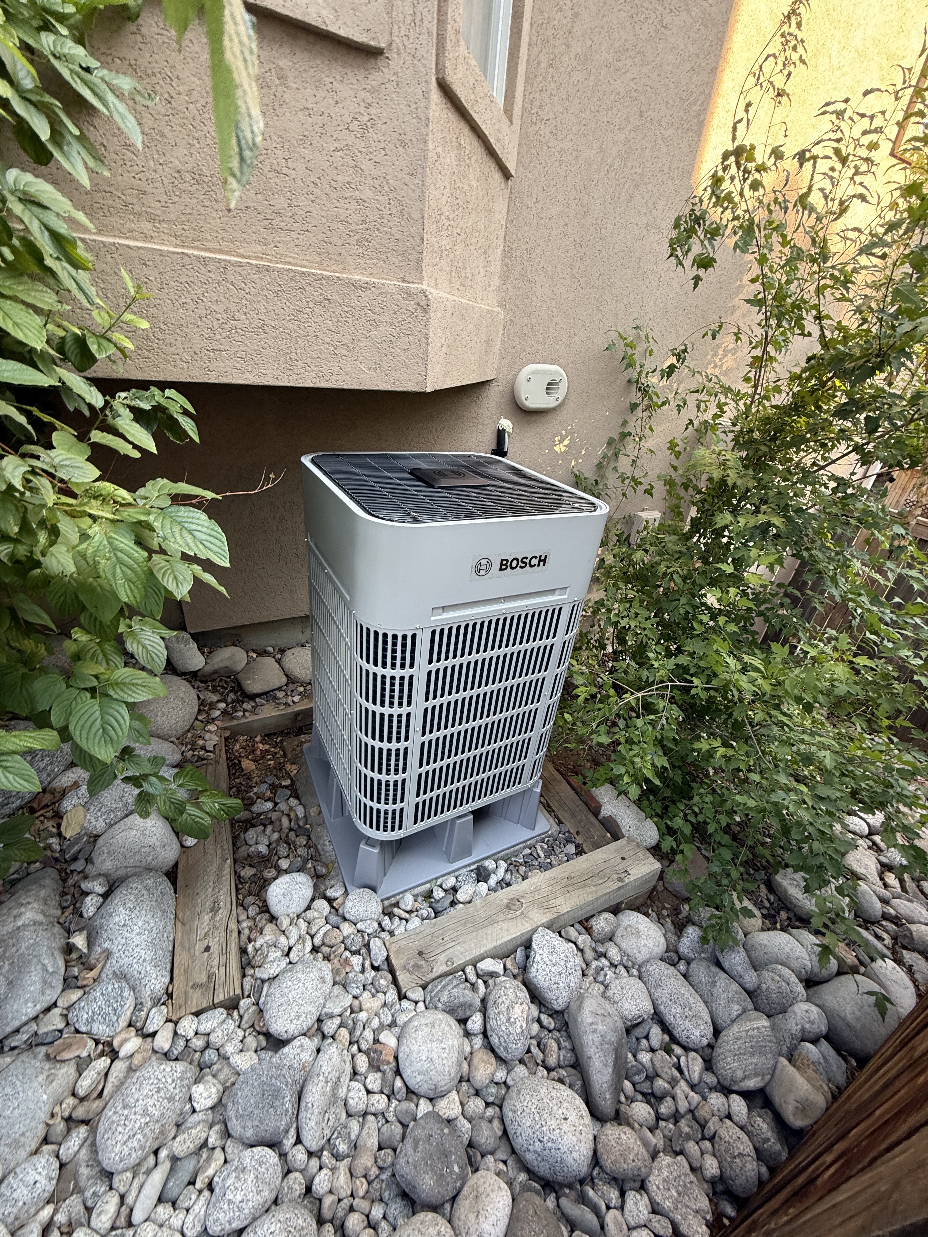 Bosch cold climate heat pump outdoor unit on a rock bed beside a stucco home